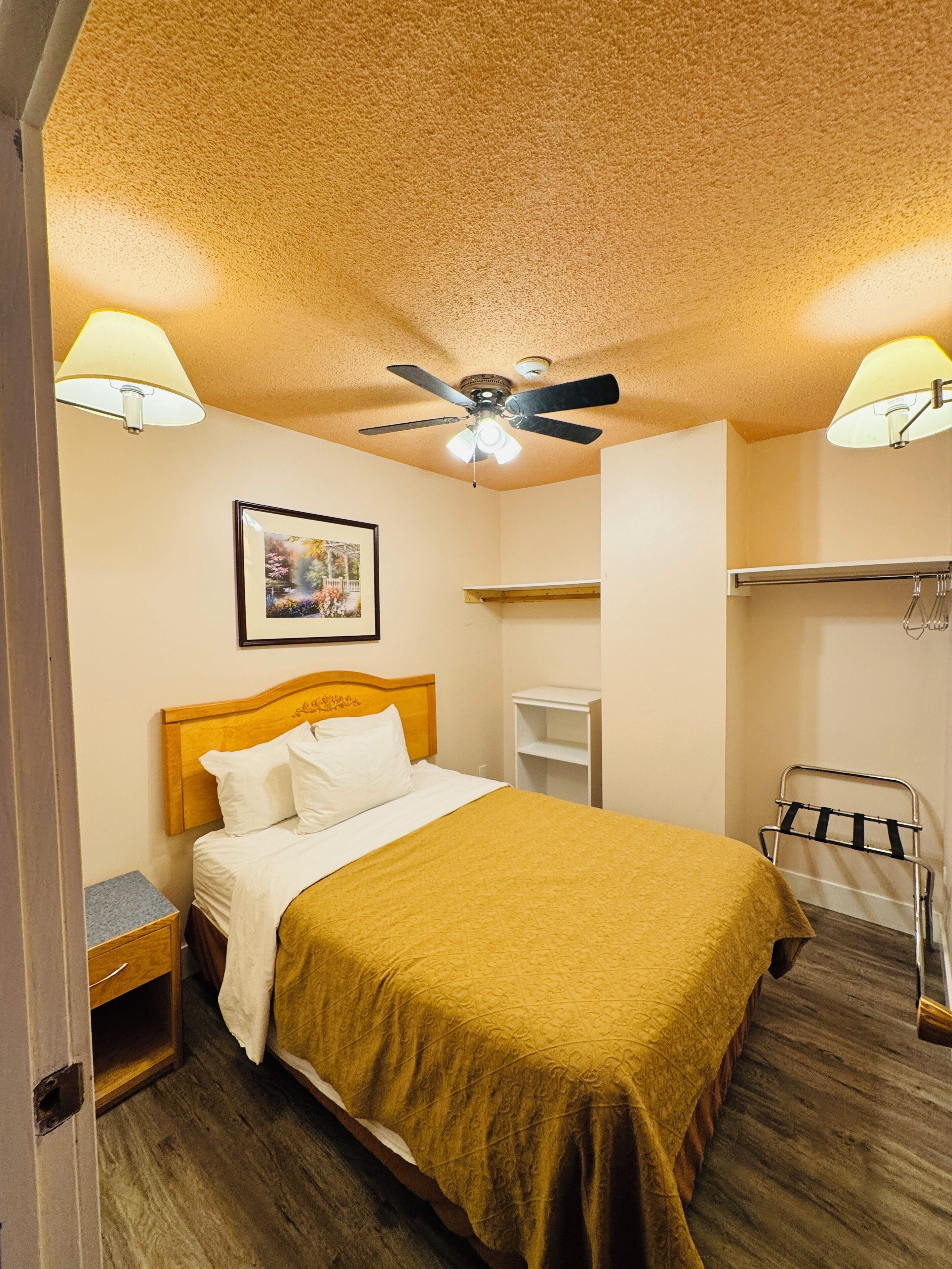 Une chambre d’hôtel équipée d’un lit, d’un ventilateur de plafond, d’une table de chevet, d’une étagère et d’un porte-serviettes.