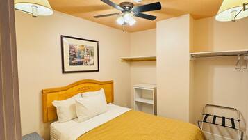 Habitación de hotel con cama, ventilador de techo, mesita de noche, estante y perchero.
