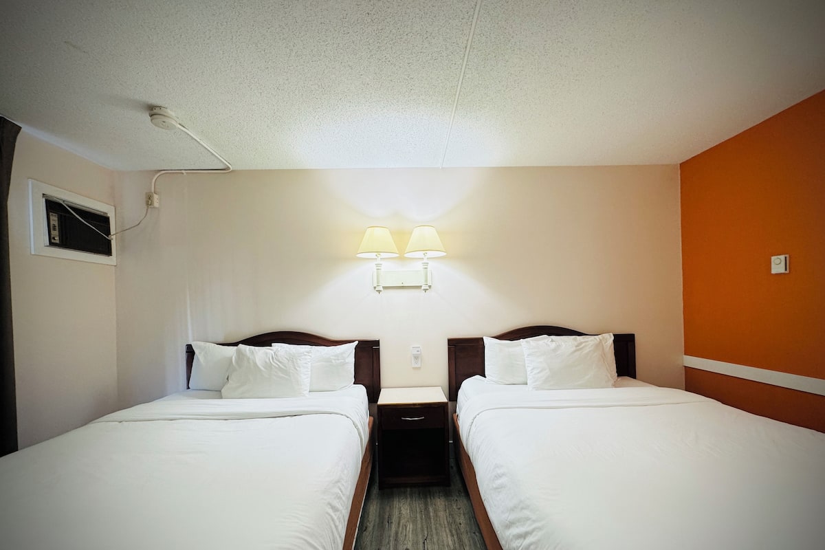 A hotel room with two single beds, a nightstand, a wall-mounted lamp, and a ceiling fan.