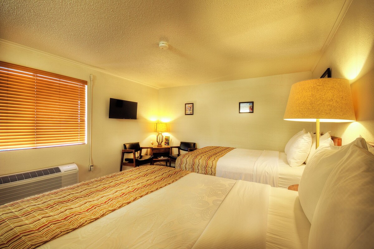 Een hotelkamer met twee bedden, een bureau met stoel, een lamp en een televisie aan de muur.