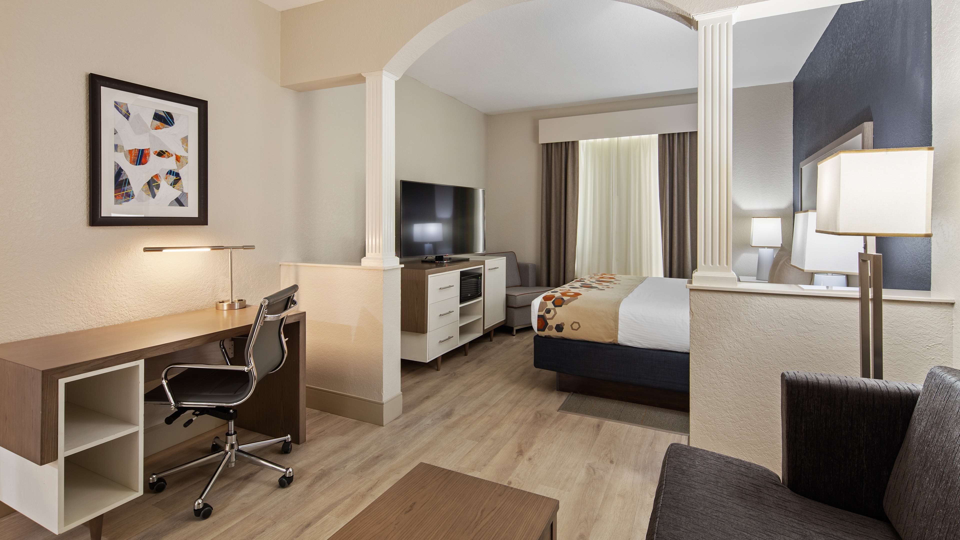 A hotel room with a desk, chair, TV, bed, and a large mirror.