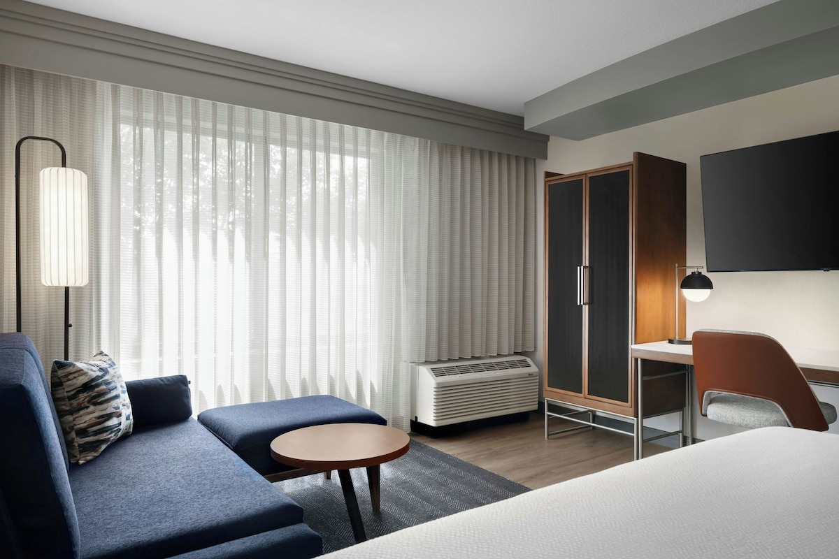 Une chambre d’hôtel avec un lit, un canapé, un bureau, une chaise, une télévision et une fenêtre avec des rideaux.