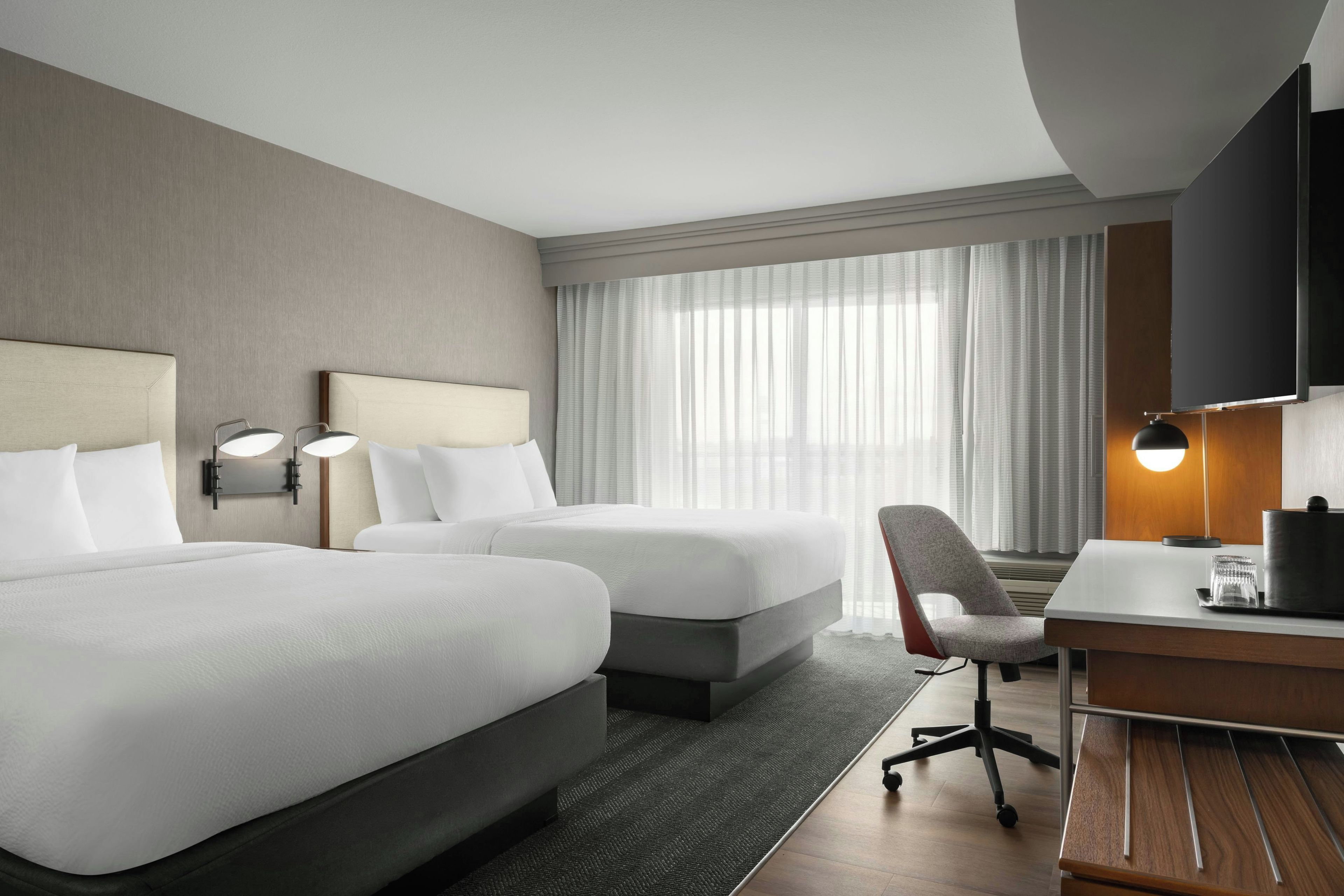 Ein Hotelzimmer mit zwei Betten, einem Schreibtisch, einem Stuhl, einem Fernseher und einem Fenster mit Vorhängen.