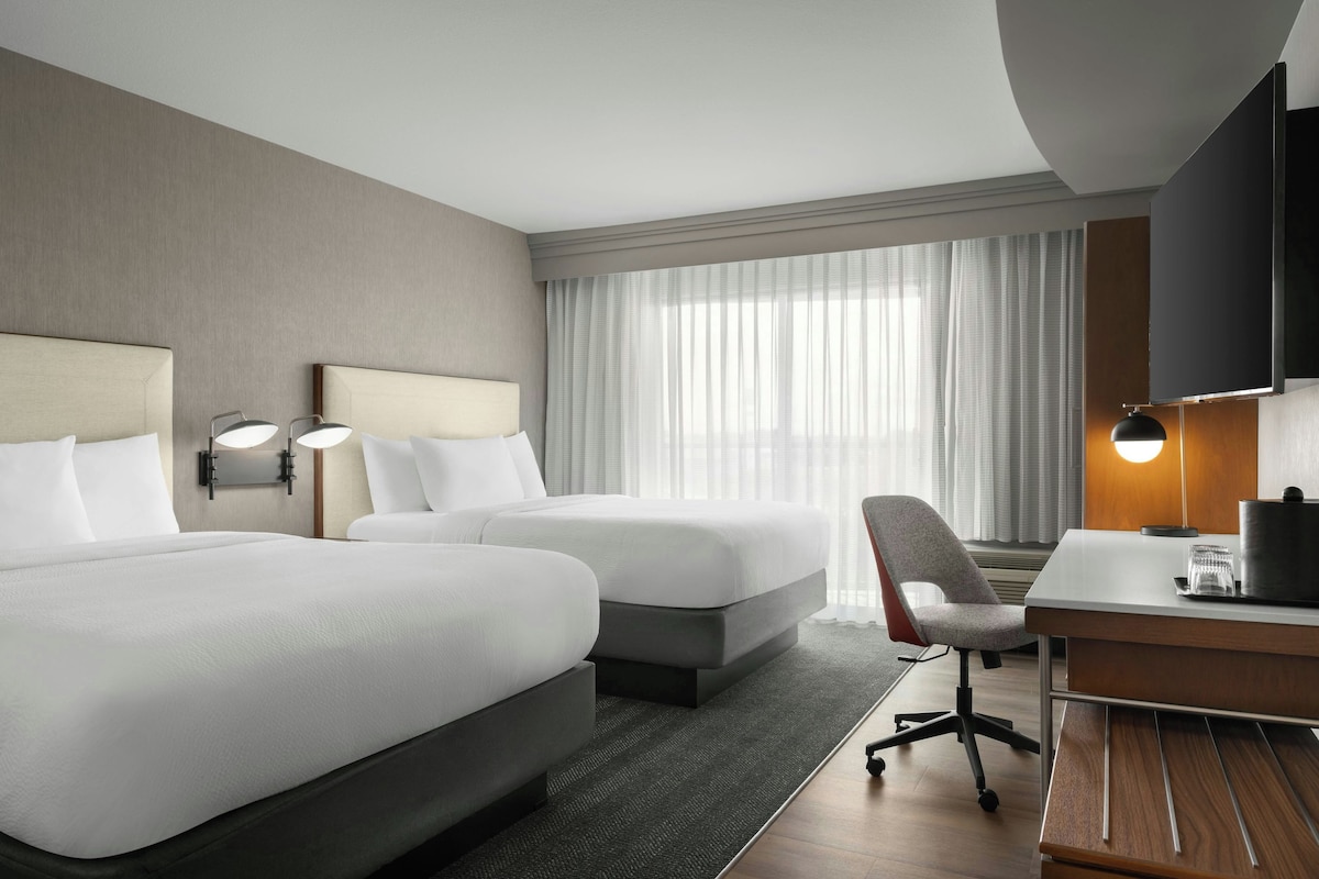 Une chambre d’hôtel avec deux lits, un bureau, une chaise, une télévision et une fenêtre avec des rideaux.