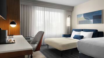 Una habitación de hotel con escritorio, una silla, una cama y un ventanal con cortinas.