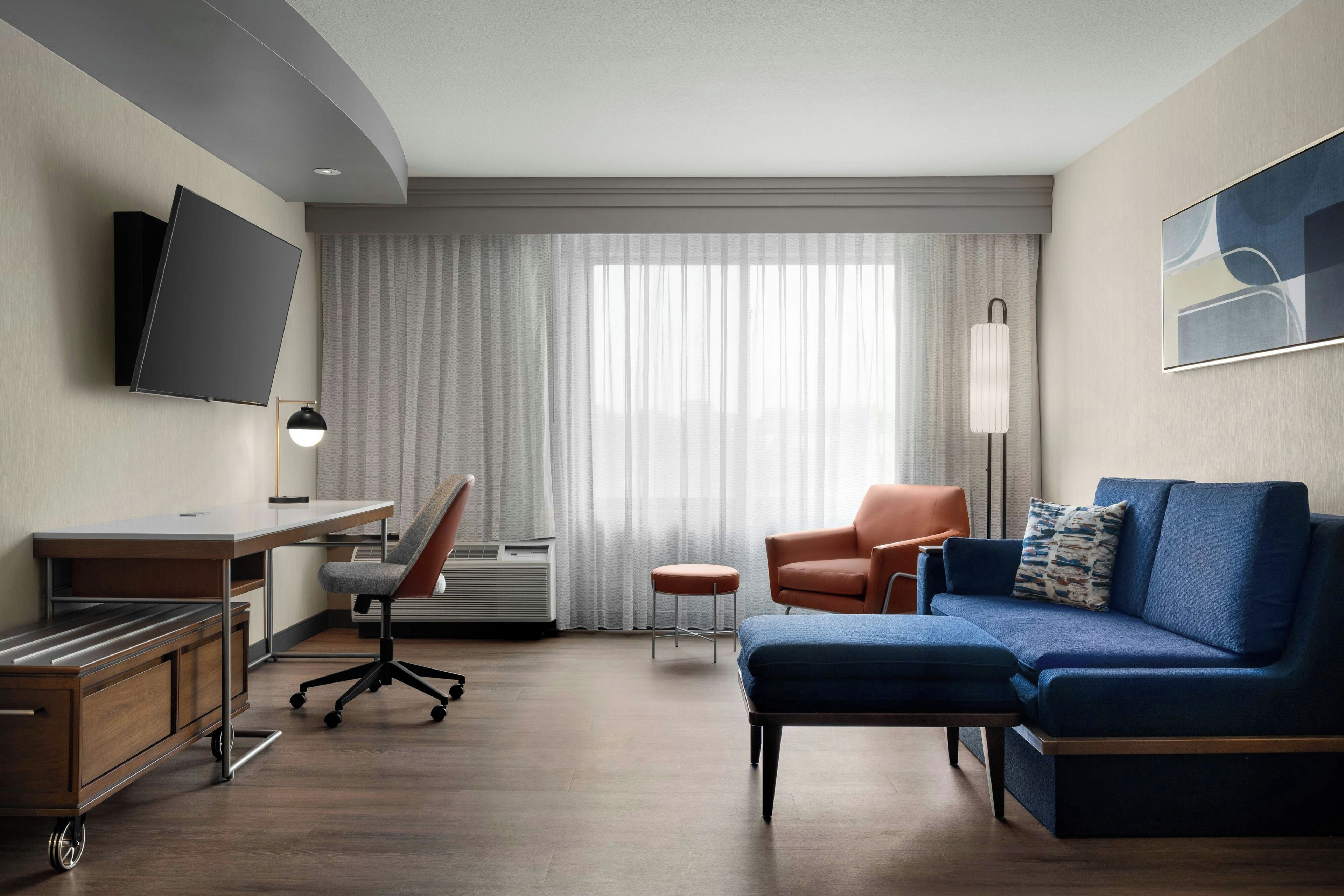 A modern hotel room with a desk, chair, sofa, TV, and a large window with curtains.