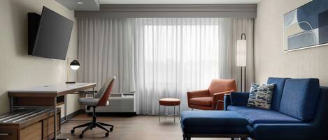 A modern hotel room with a desk, chair, sofa, TV, and a large window with curtains.