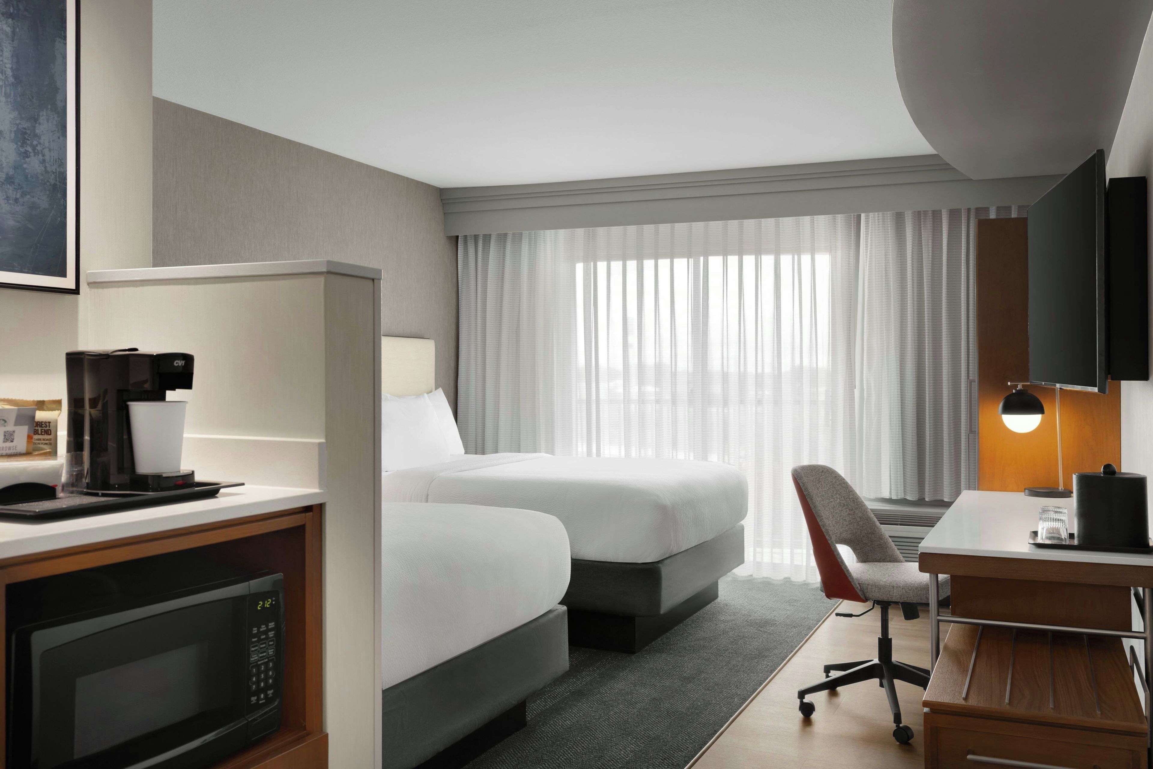 Ein Hotelzimmer mit Bett, Schreibtisch inklusive Kaffeemaschine, Stuhl, Fernseher und einem Fenster mit Vorhängen.