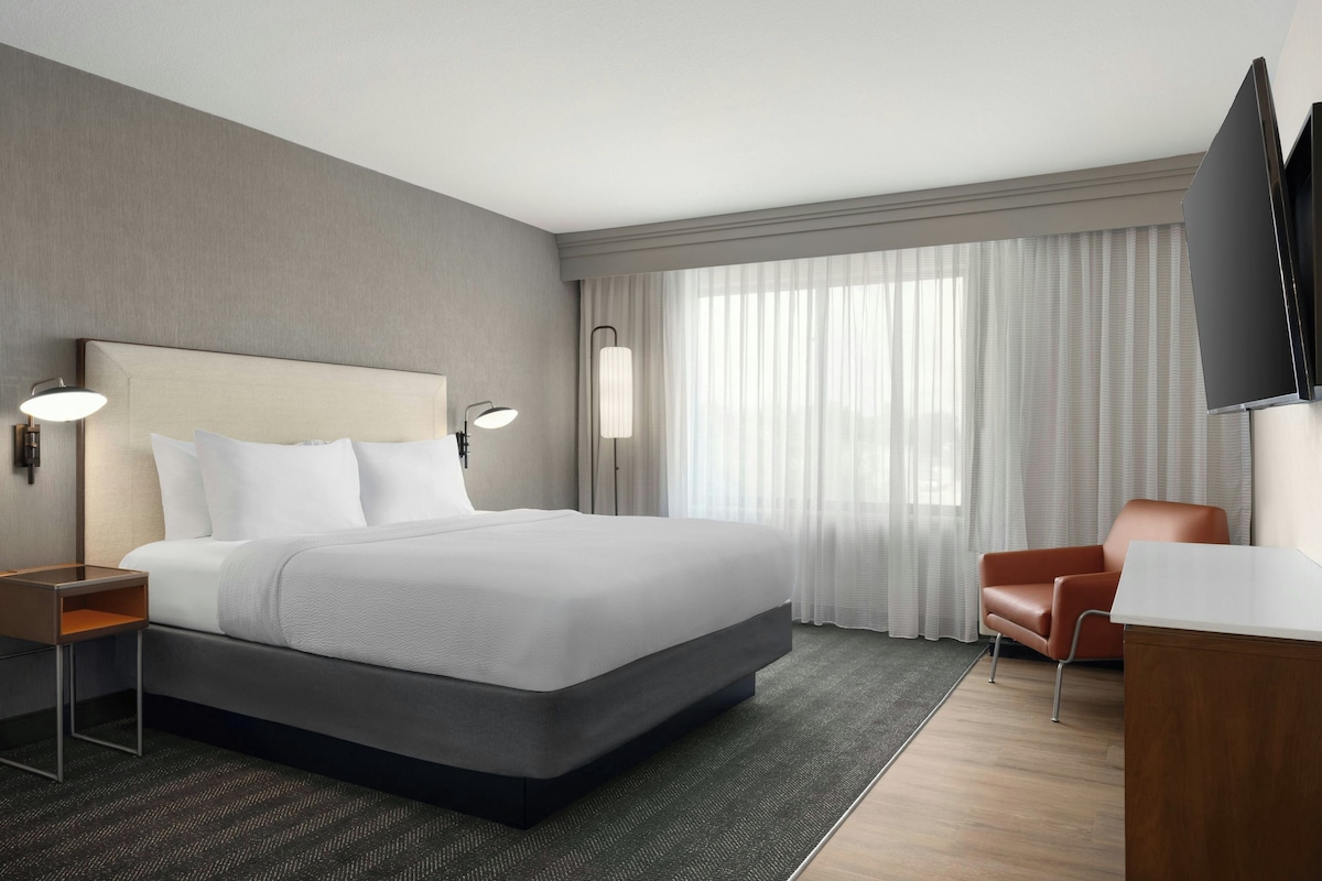 Une chambre d’hôtel avec un grand lit, un bureau, une chaise, une télévision et une fenêtre avec des rideaux.