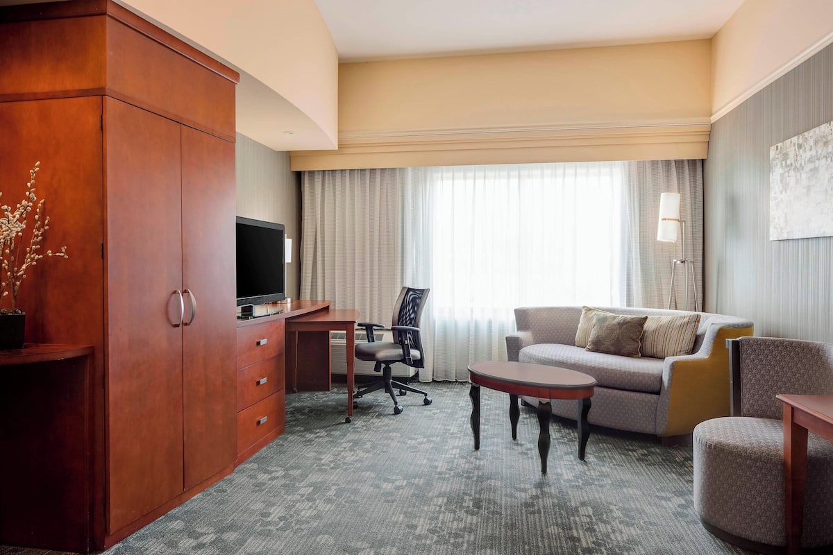 Ett hotellrum med en garderob i trä, ett skrivbord med en tv, en soffa, ett soffbord och en stol.