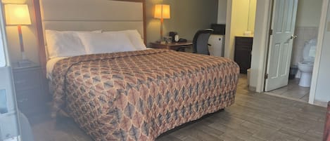 Une chambre d’hôtel avec un grand lit, deux lampes de chevet, un bureau avec une chaise, un petit réfrigérateur et une porte donnant sur une salle de bain.