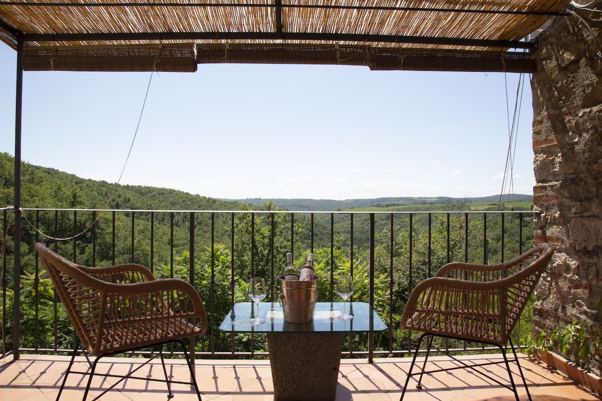 A balcony with wicker chairs, a glass table, and a view of a lush green landscape.