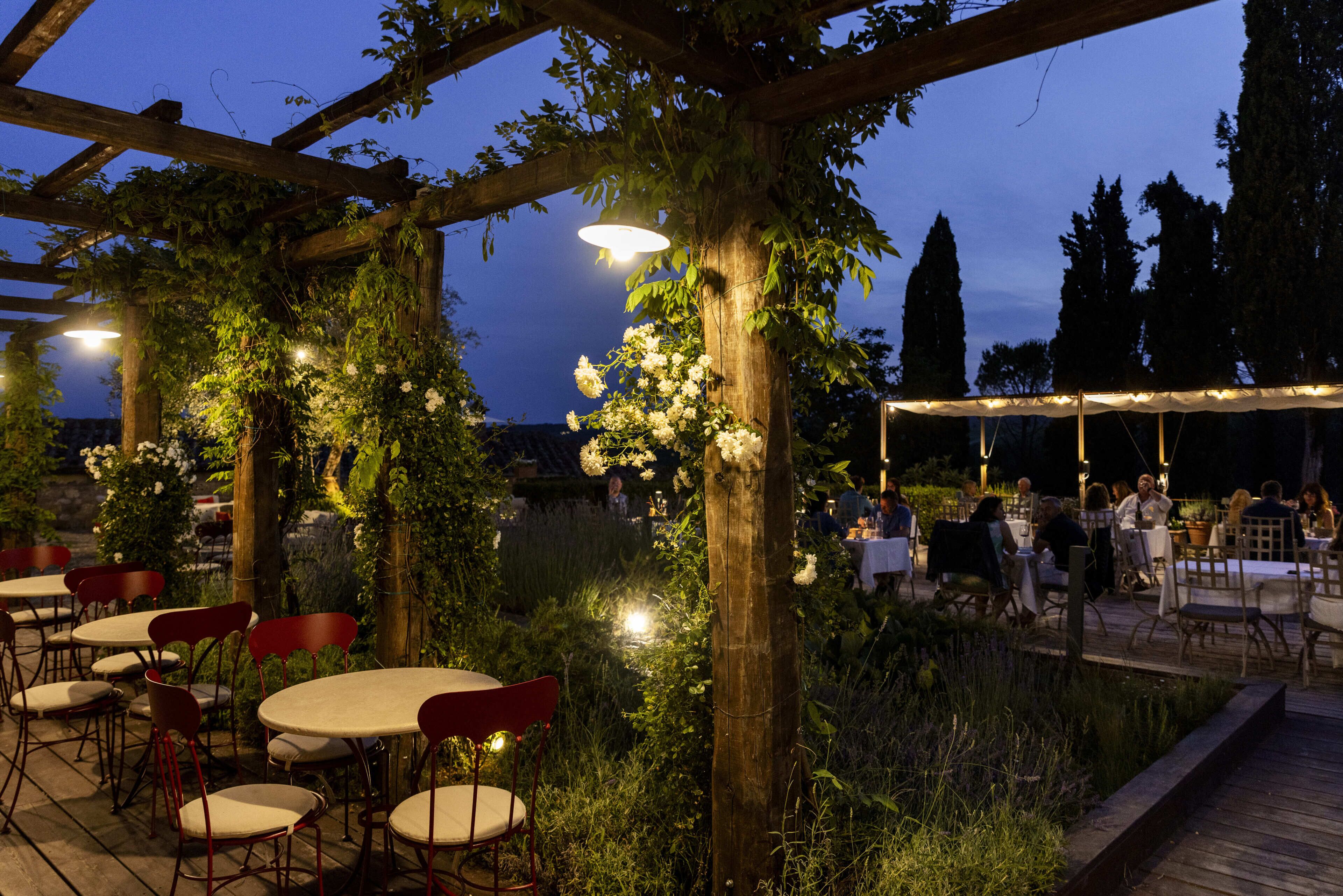 An outdoor dining area with tables and chairs, surrounded by greenery and under a pergola structure.