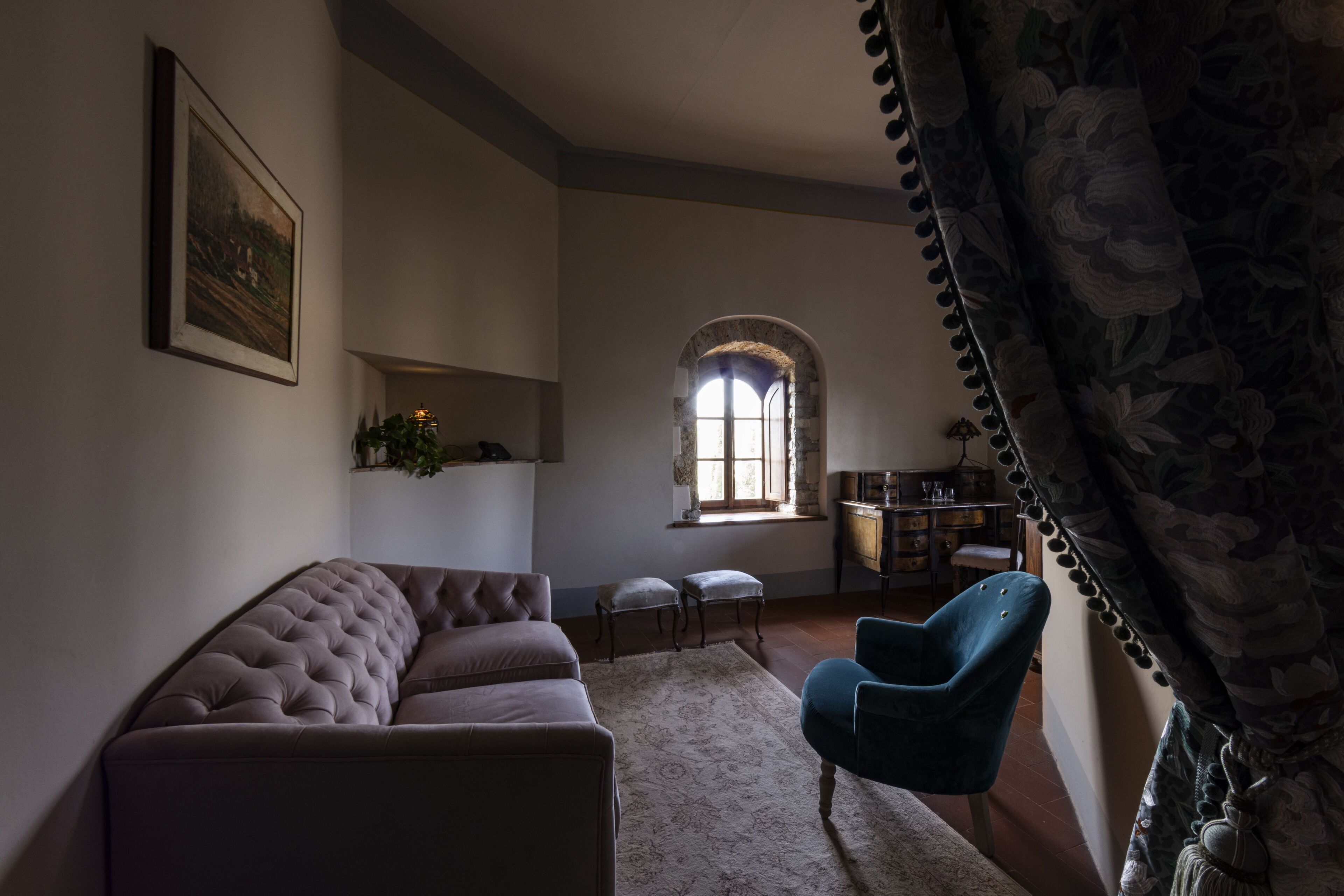Ein Wohnzimmer mit gepolstertem Sofa, blauem Sessel, einem gemusterten Teppich, einem gerahmten Bild und einem Fenster mit rundbogigem Steinrahmen.