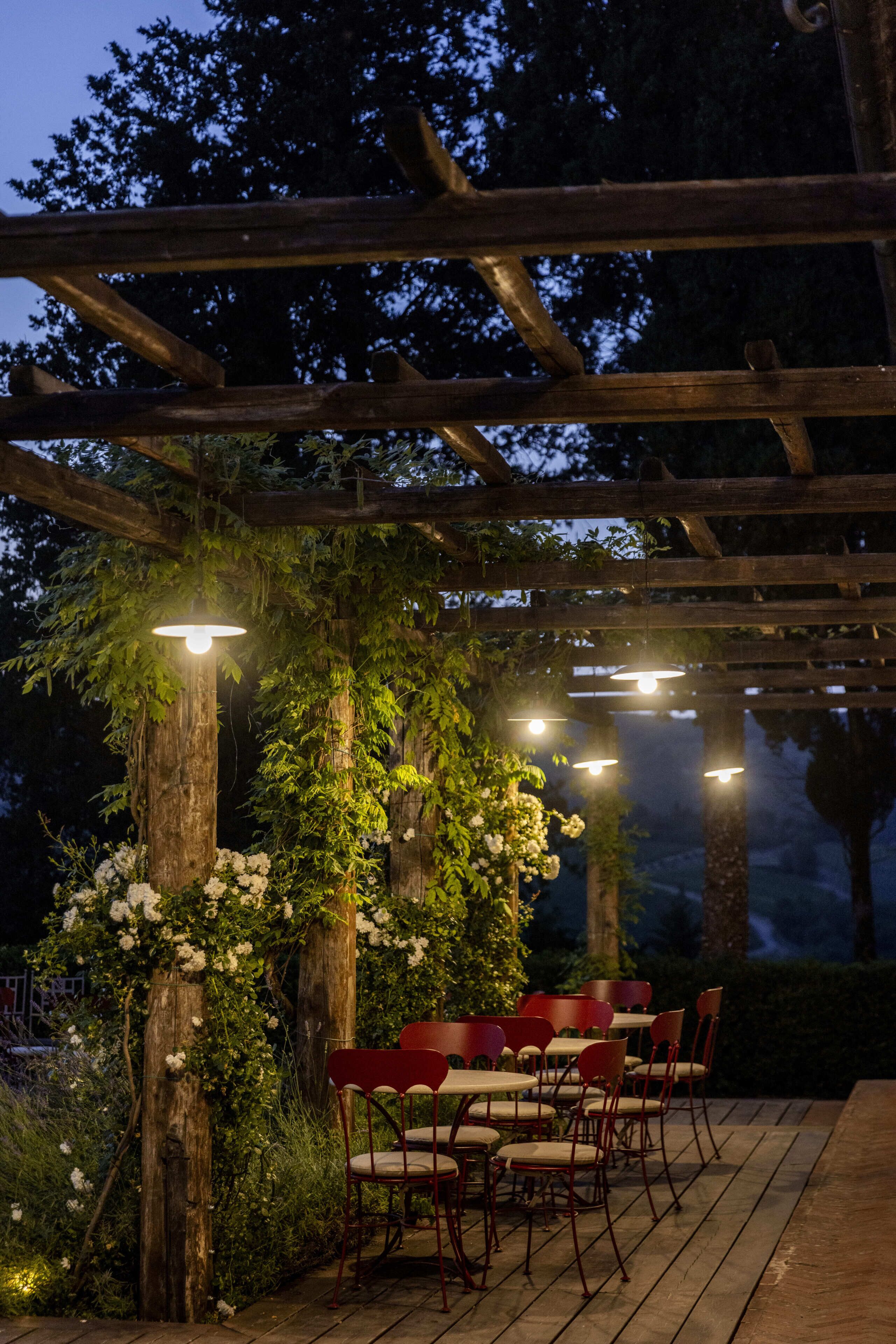 Una pergola in legno con tavoli e sedie sotto, circondata da piante e fiori.