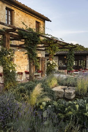 Un edificio rústico de piedra con una pérgola de madera, mobiliario exterior y un jardín con diversas plantas.