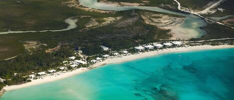 An aerial view of a coastal resort with multiple buildings along the beachfront, surrounded by natural landscapes and clear turquoise waters.