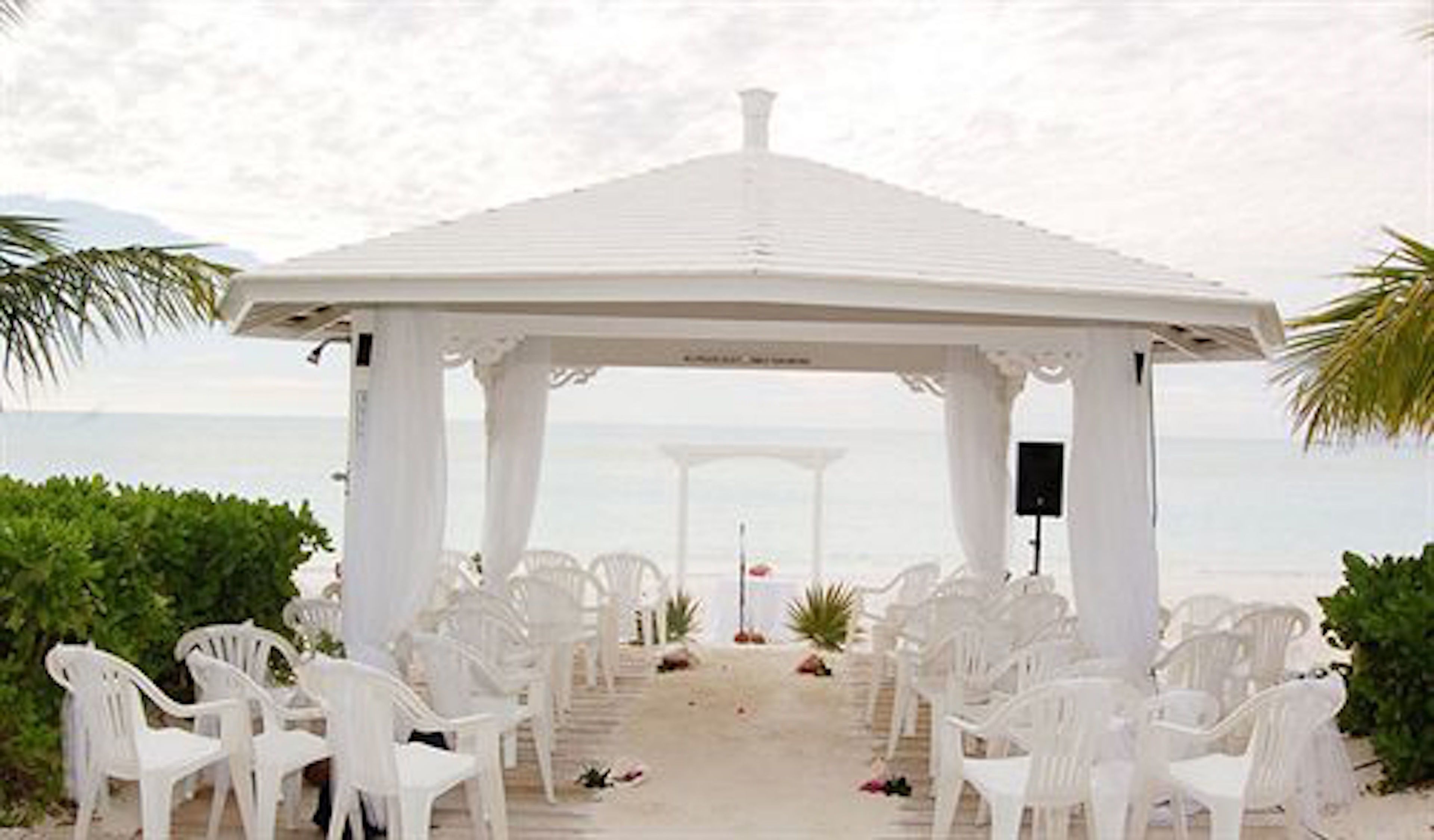 A white gazebo with a tiled roof, surrounded by white plastic chairs, set on a sandy beach with a view of the sea.