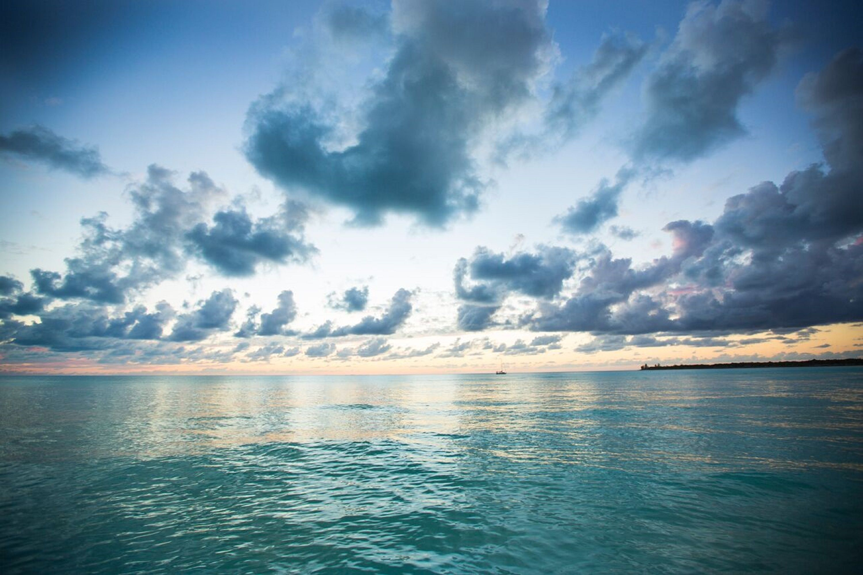 Une mer calme, avec un navire au loin et une rangée de bâtiments à l’horizon, sous un ciel partiellement nuageux.