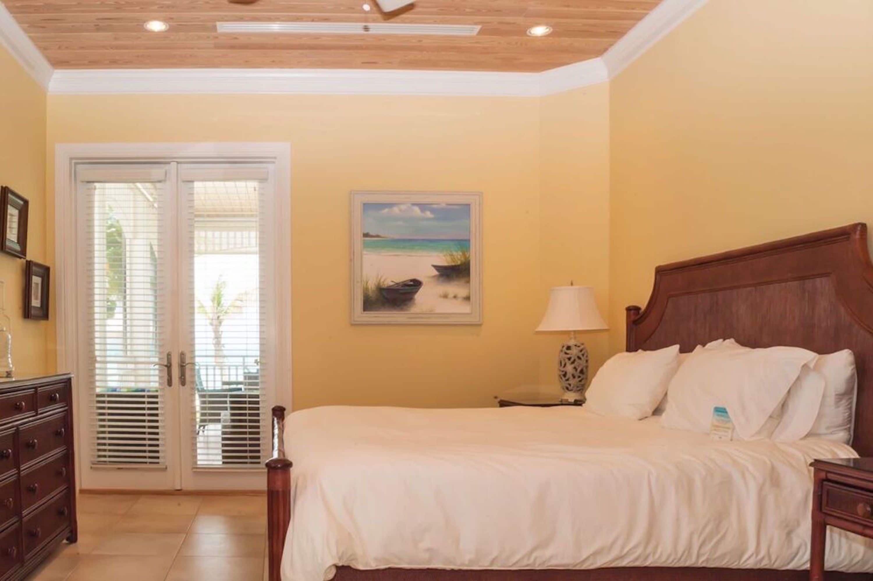 A bedroom with a bed, a nightstand, a lamp, a painting, and a door with blinds.