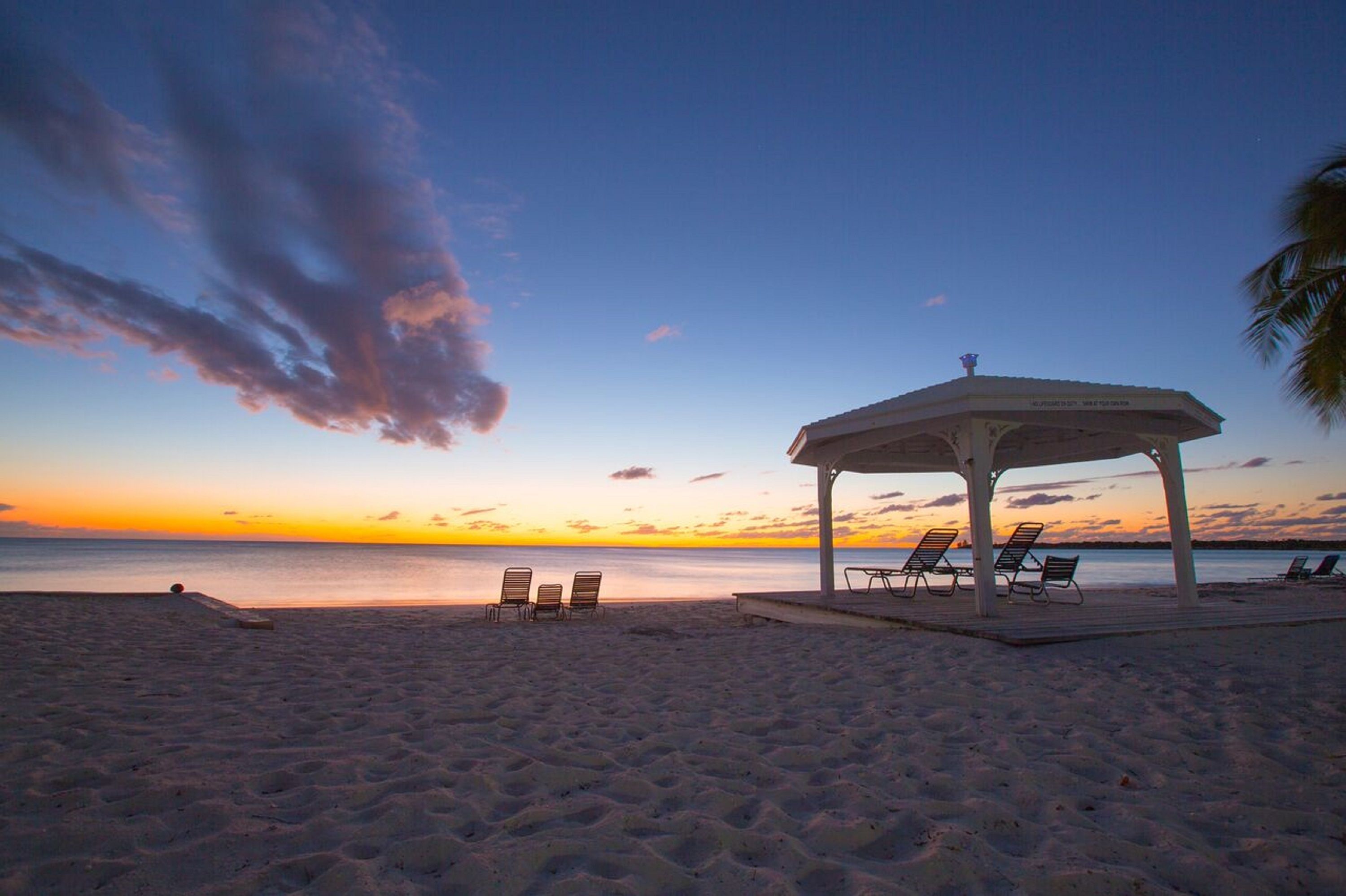 A beach with a gazebo, lounge chairs, and a calm sea at sunset.