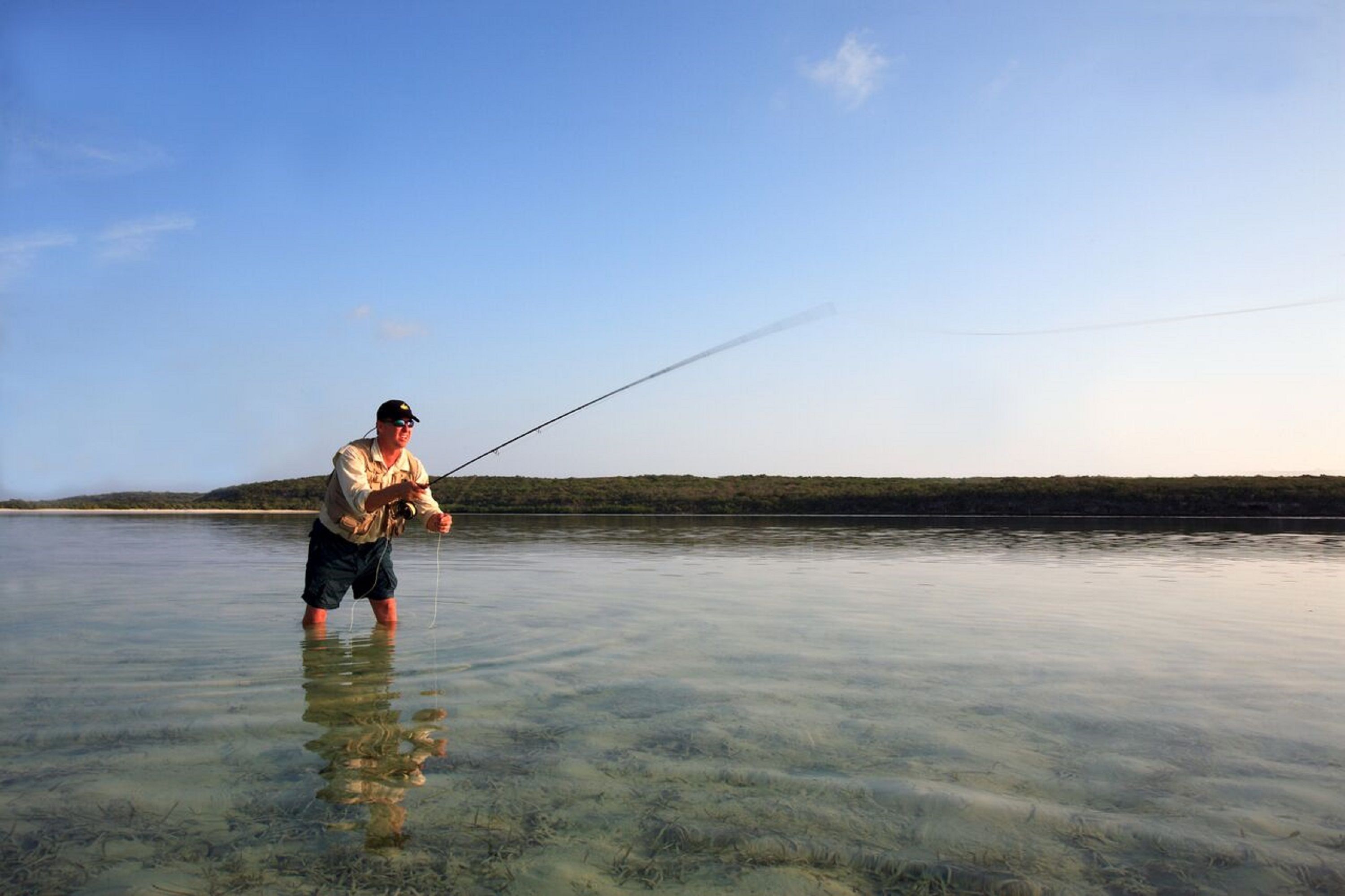 Une personne est en train de pêcher dans une eau claire et peu profonde, avec une côte visible au loin.