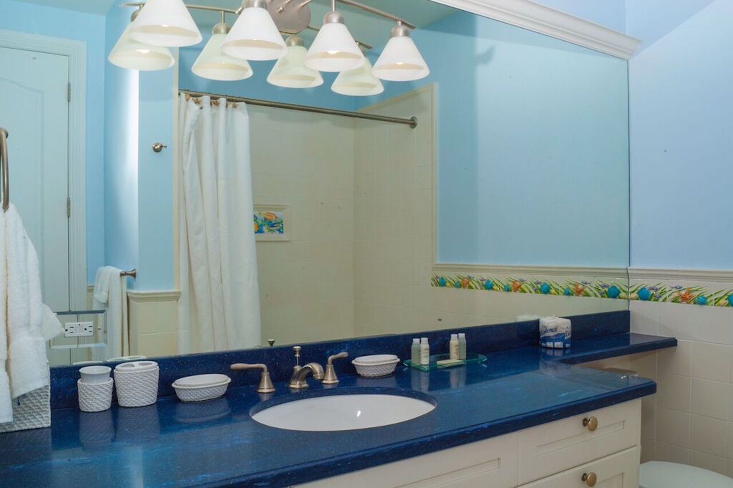 A bathroom with a blue countertop, white sink, and a large mirror. There are multiple light fixtures above the mirror and a shower curtain visible in the background.