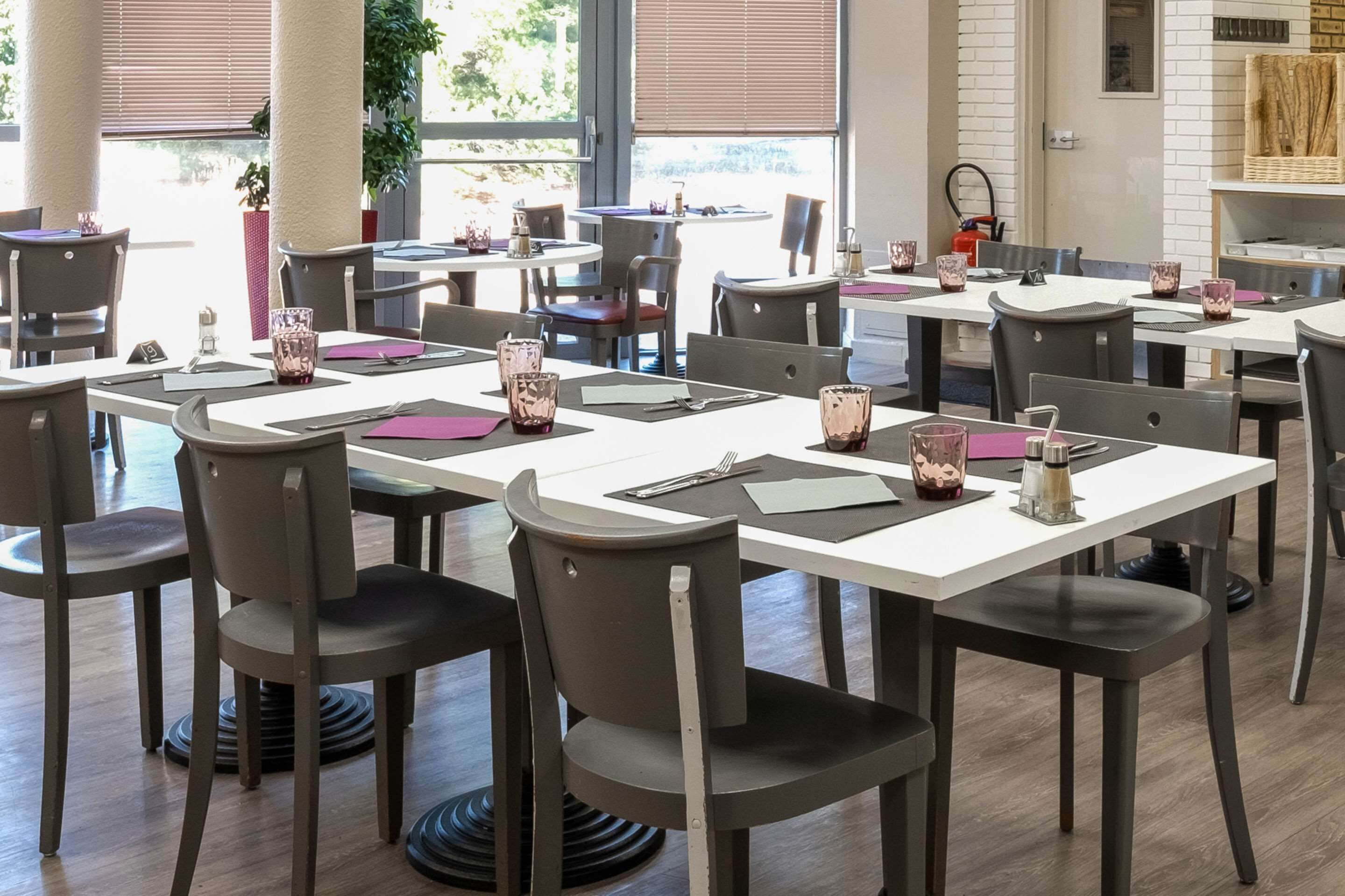Un restaurante con varias mesas y sillas, dispuesto en un espacio amplio con luz natural que entra por grandes ventanales.