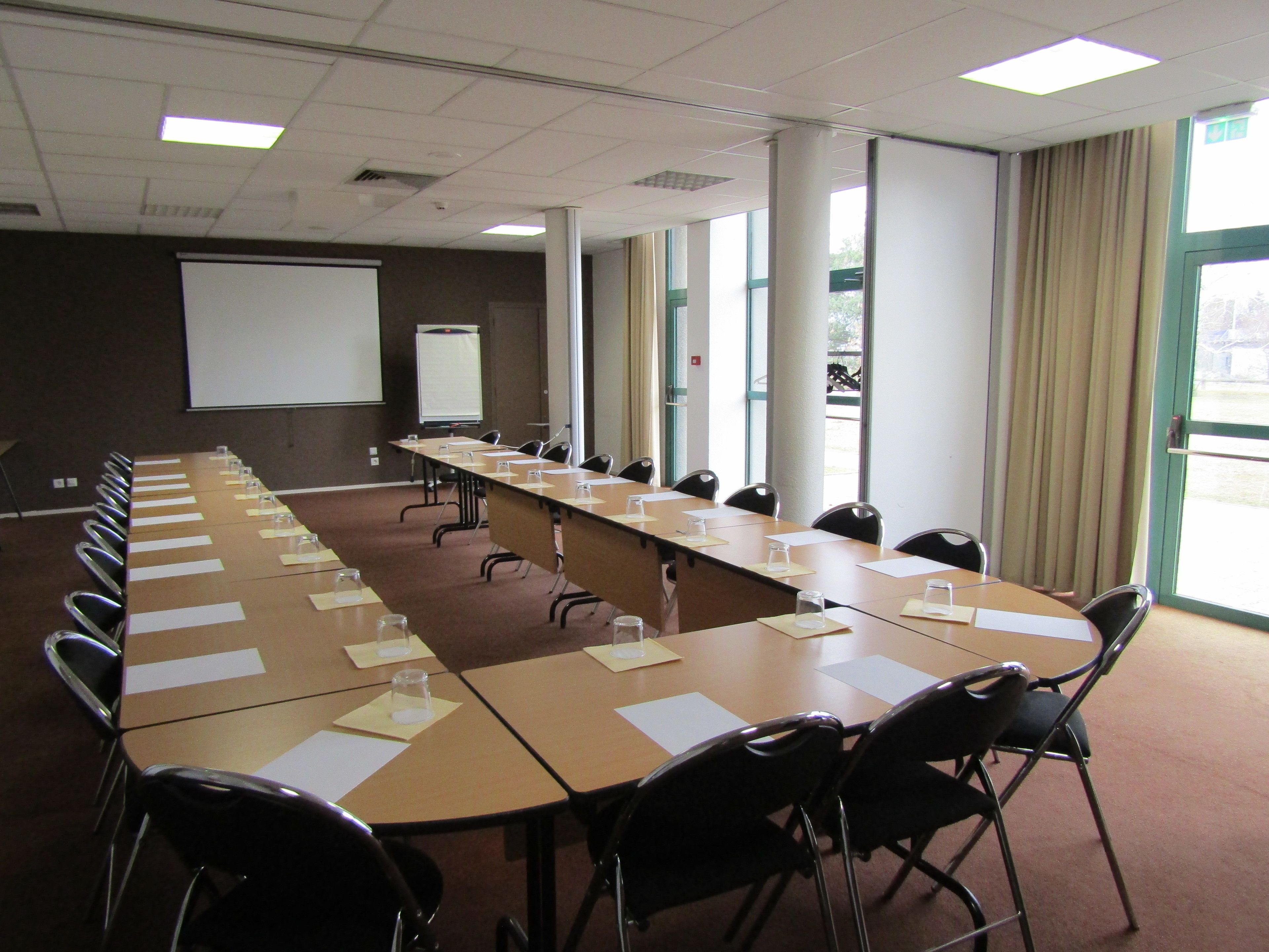 Un salón de conferencias con una mesa larga, sillas, una pantalla de proyector y ventanas amplias.