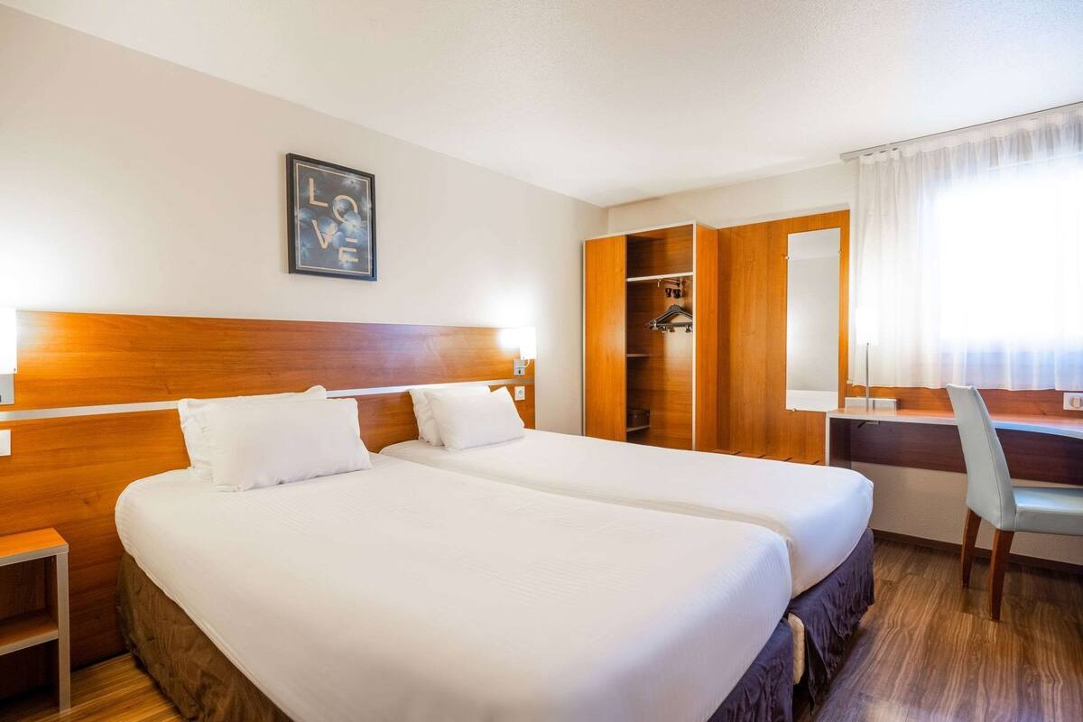 Habitación de hotel con dos camas, un escritorio, una silla, un armario y un cuadro en la pared.