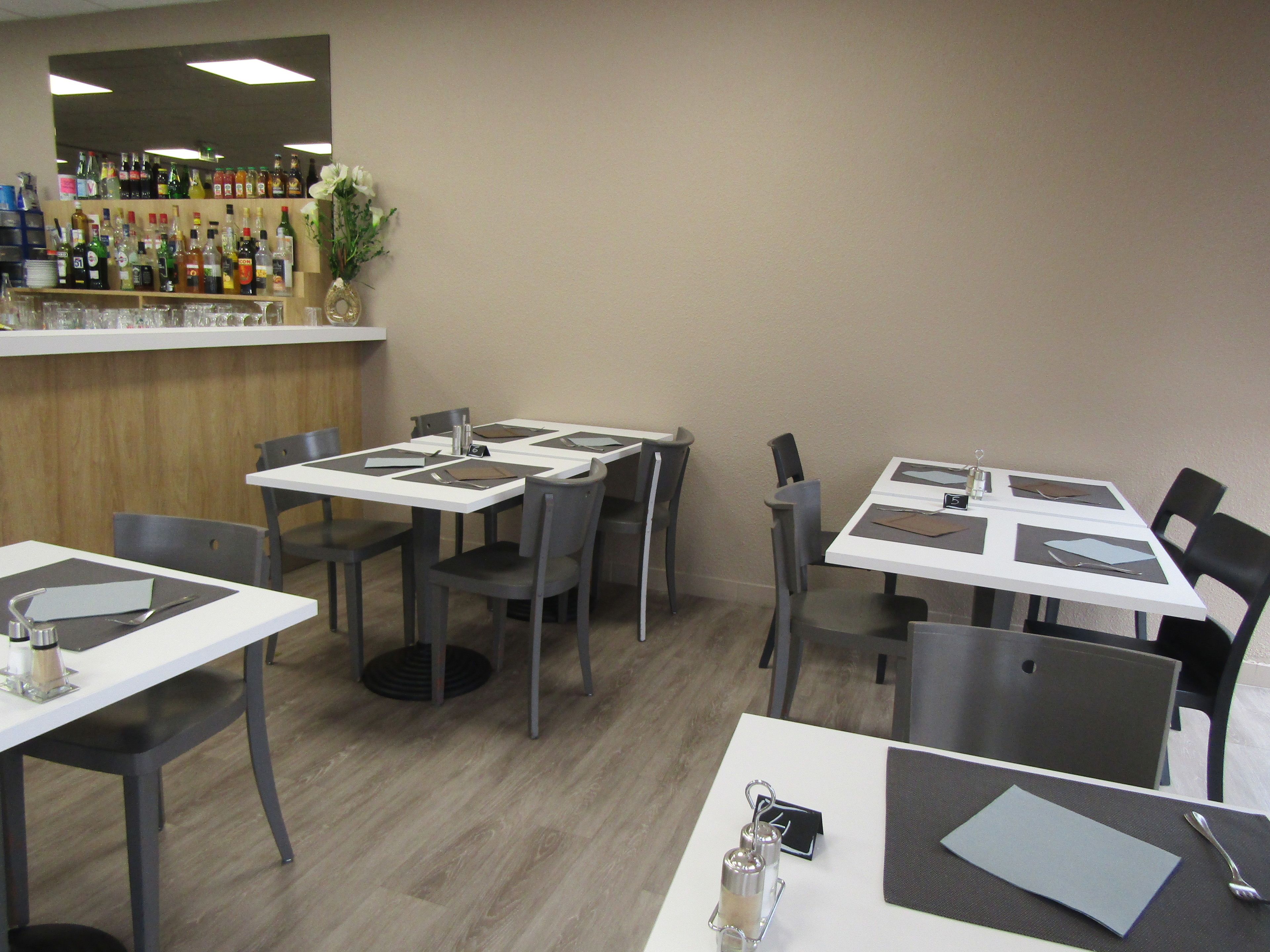 A restaurant interior with tables set for dining, chairs, a bar area with various bottles, and a vase with flowers.