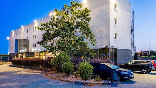 A modern hotel with a glass facade, a tree in front, and a parking lot with cars.
