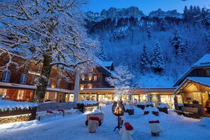 A snow-covered courtyard with a wooden building, a snow-covered tree, and a mountain backdrop. - Kemmeriboden Bad Swiss Quality Hotel (Schangnau)