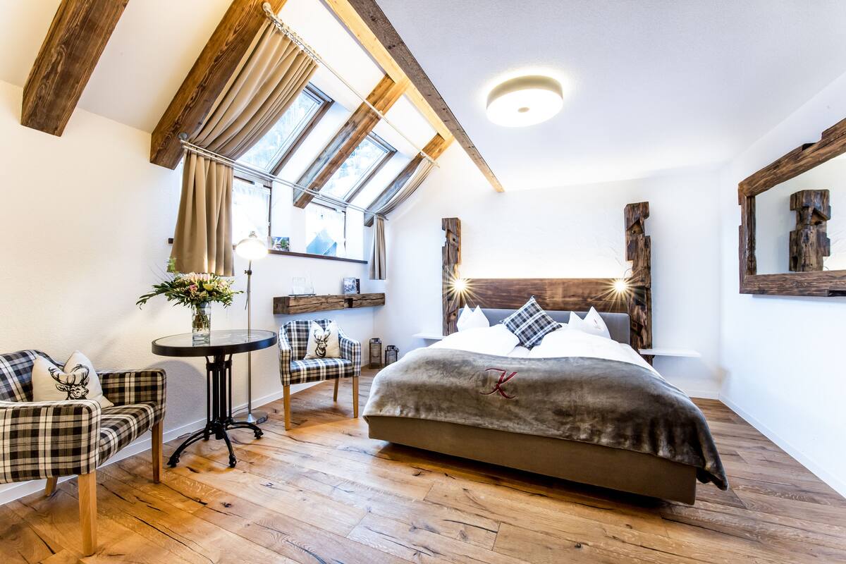 Ein Schlafzimmer mit einem großen Bett, einem kleinen Tisch, einem Sessel und einer Couch. Es gibt Fenster mit Vorhängen, Holzbalken an der Decke und einen Spiegel an der Wand.