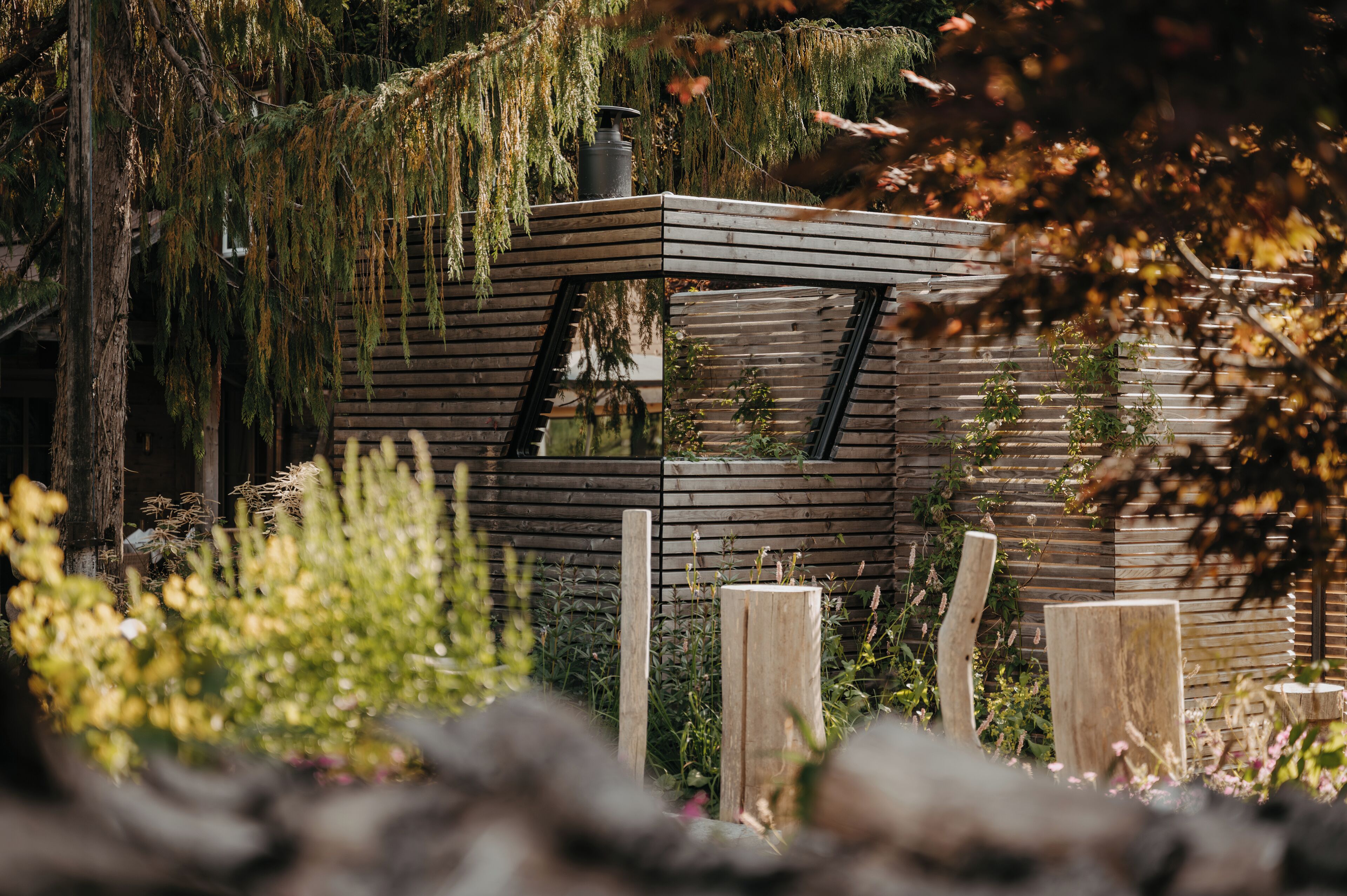 Een moderne, meerlaagse houten constructie met een gemêleerd dak, omgeven door weelderige begroeiing en bomen.
