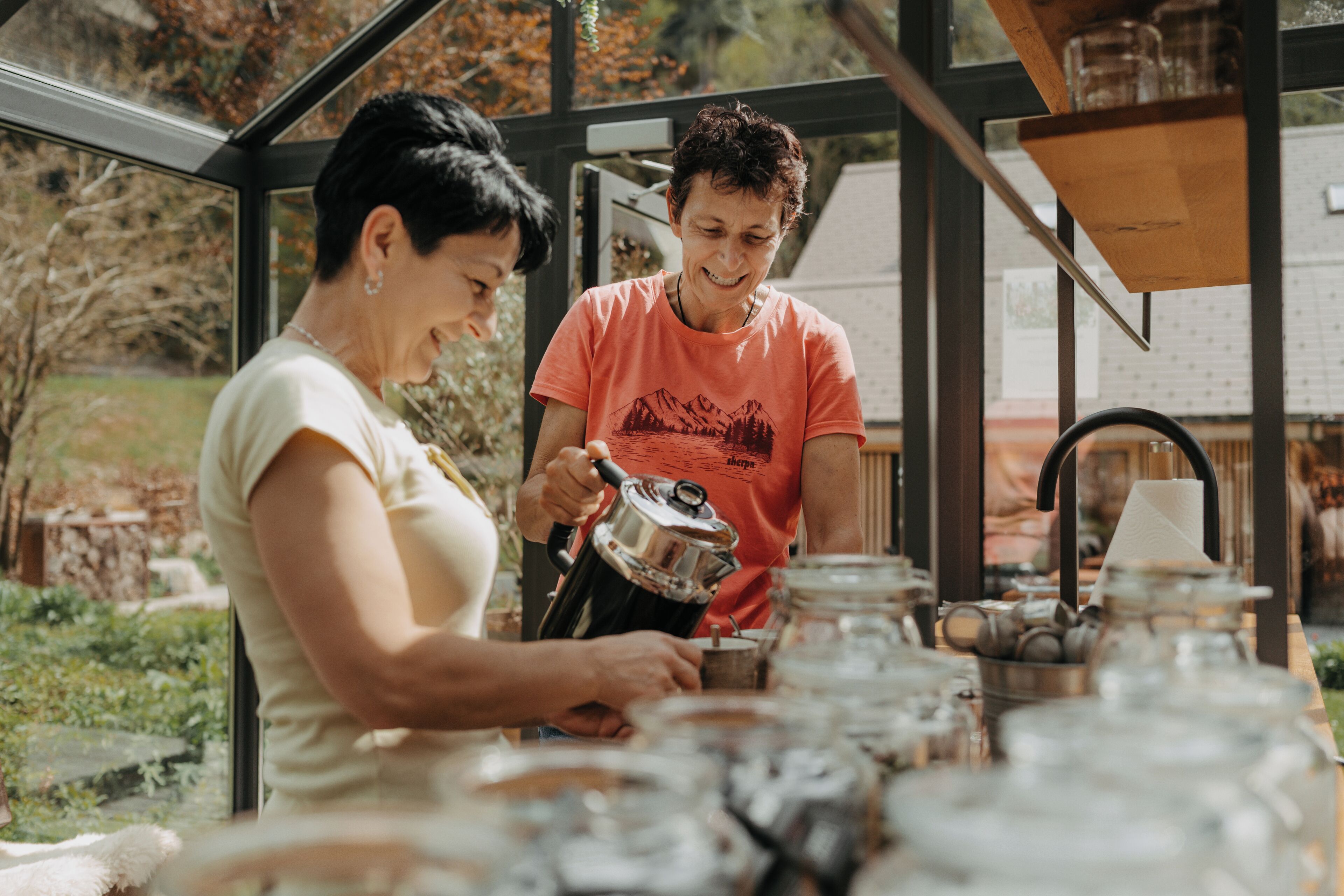 Dos personas preparando café en una cocina con un ventanal grande, un estante de madera y varios utensilios de cocina.
