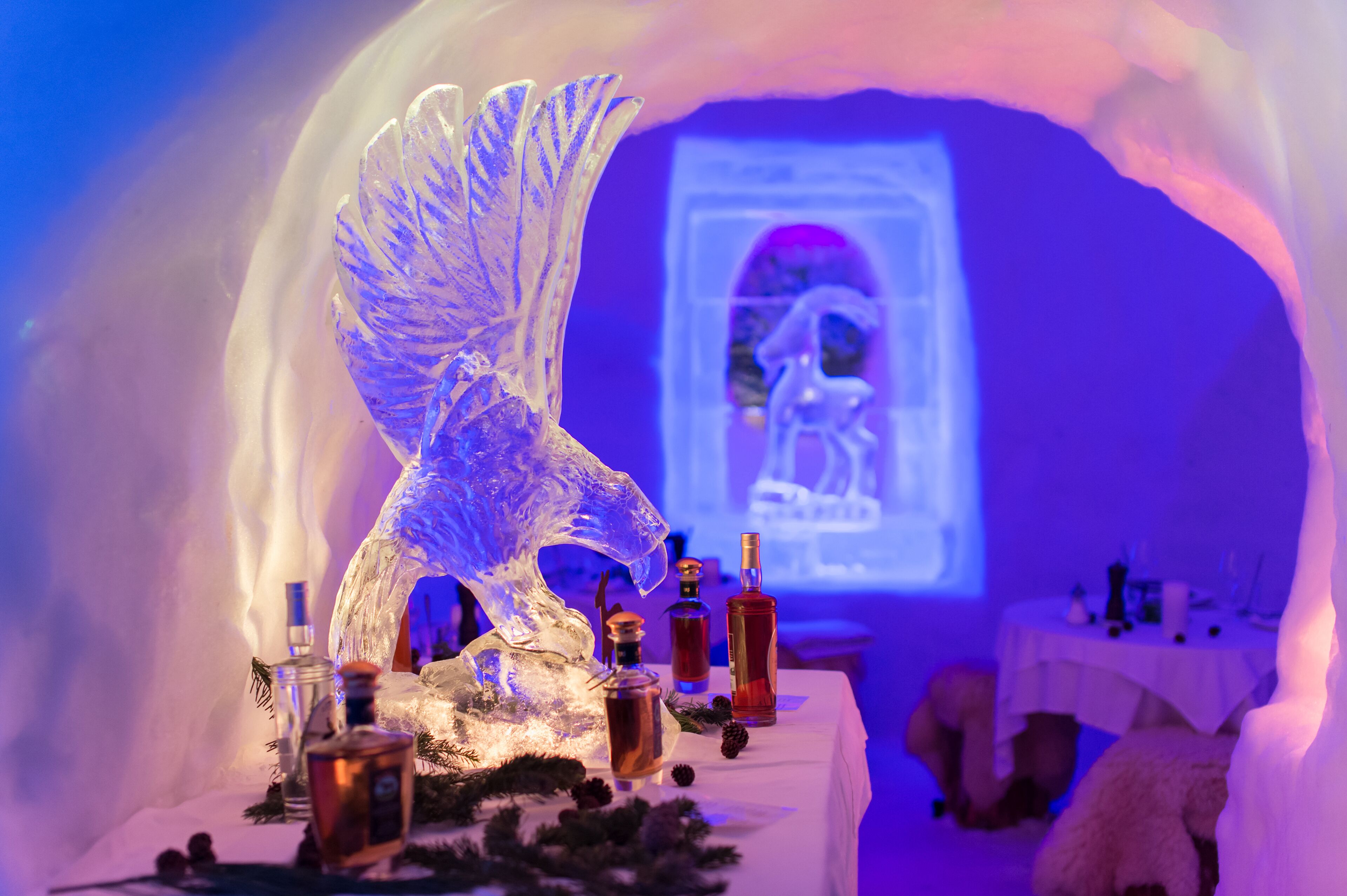 Una escultura de hielo de una criatura alada, una proyección de una persona y varias botellas sobre una mesa en un túnel de hielo.