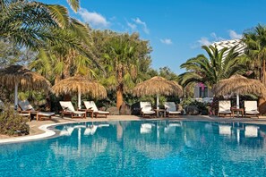 A swimming pool with lounge chairs and thatched umbrellas, surrounded by palm trees and a clear sky. - Atlantis Beach Villa (Santorini)