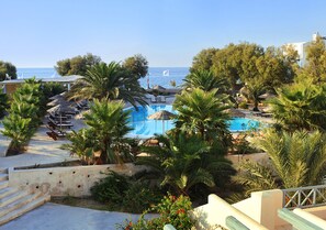 A pool area with lounge chairs and umbrellas, surrounded by palm trees and a view of the sea. - Atlantis Beach Villa (Santorini)