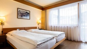 A hotel room with two beds, wooden headboards, a framed picture on the wall, and a large window with sheer curtains.