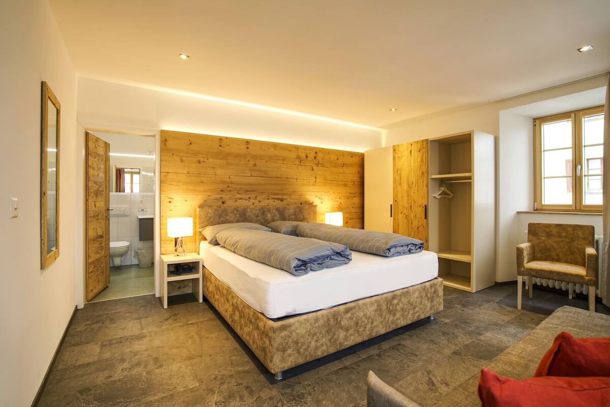 A modern bedroom with a wooden headboard, a large bed, bedside tables with lamps, a wardrobe, and a chair.