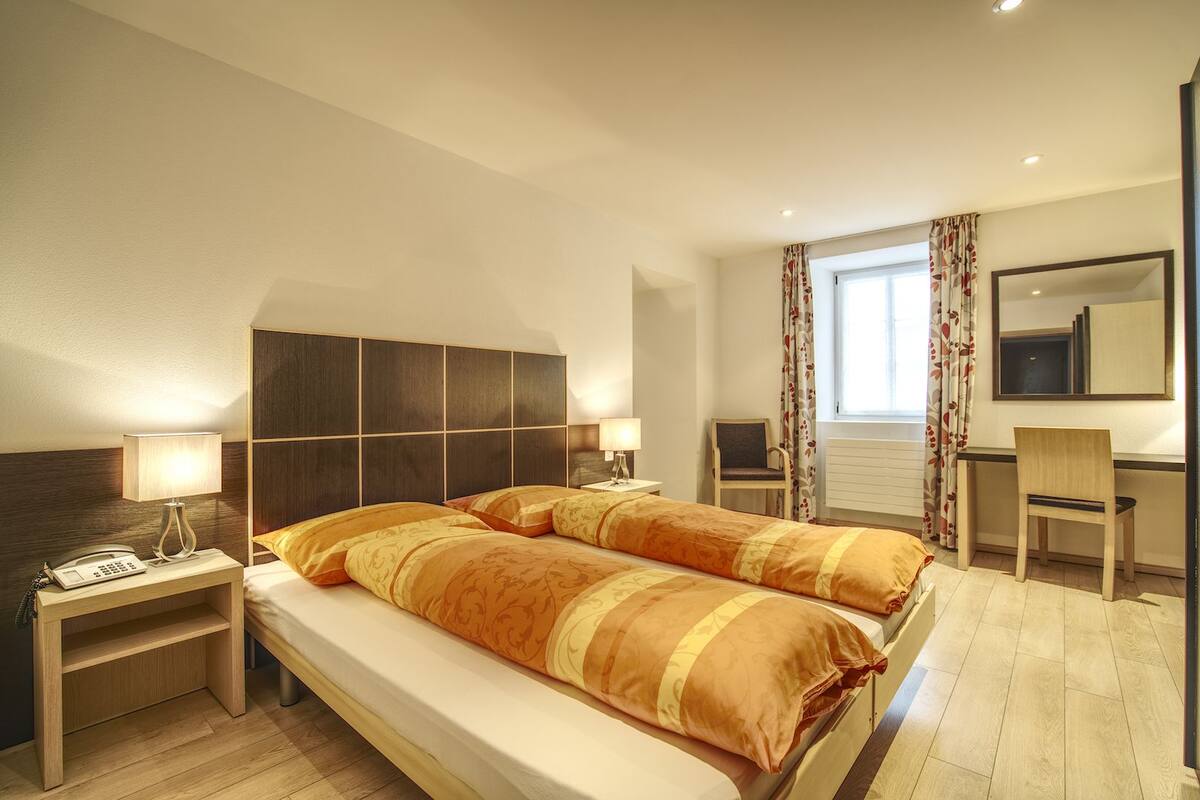 A hotel room with a large bed, bedside tables with lamps, a desk with a chair, a window with curtains, and a mirror on the wall.
