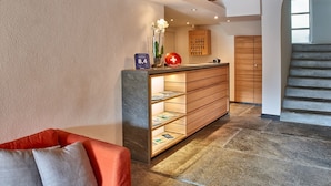 A hotel reception area with a wooden reception desk, a glass vase with flowers, a red hotel sign, and a staircase.