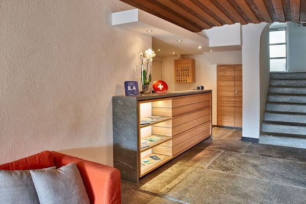 Zona de recepción de hotel con un mostrador de madera, un jarrón de cristal con flores, un letrero rojo del hotel y una escalera.