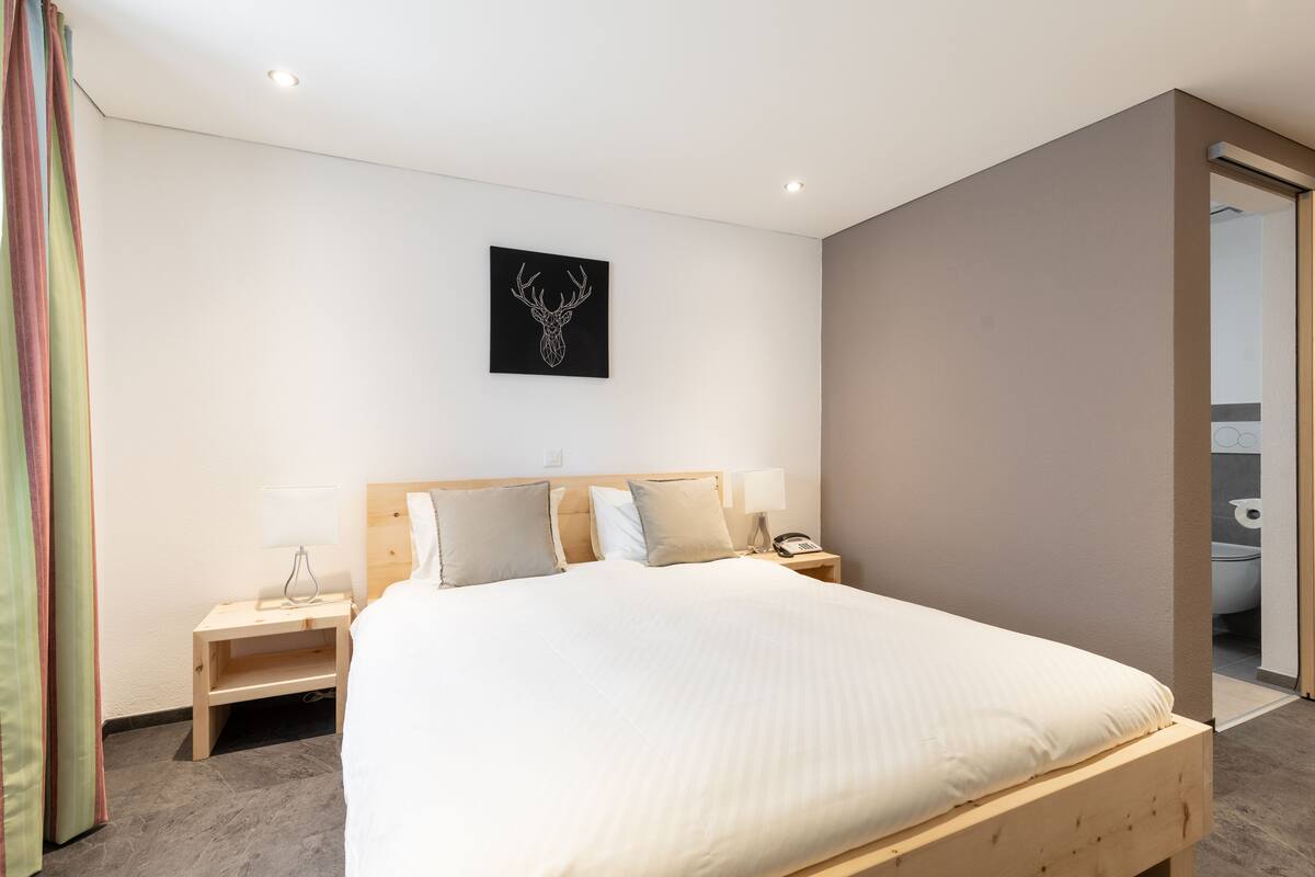 A modern hotel room with a wooden bed, a bedside table with a lamp, a wall-mounted phone, and a framed deer head artwork.