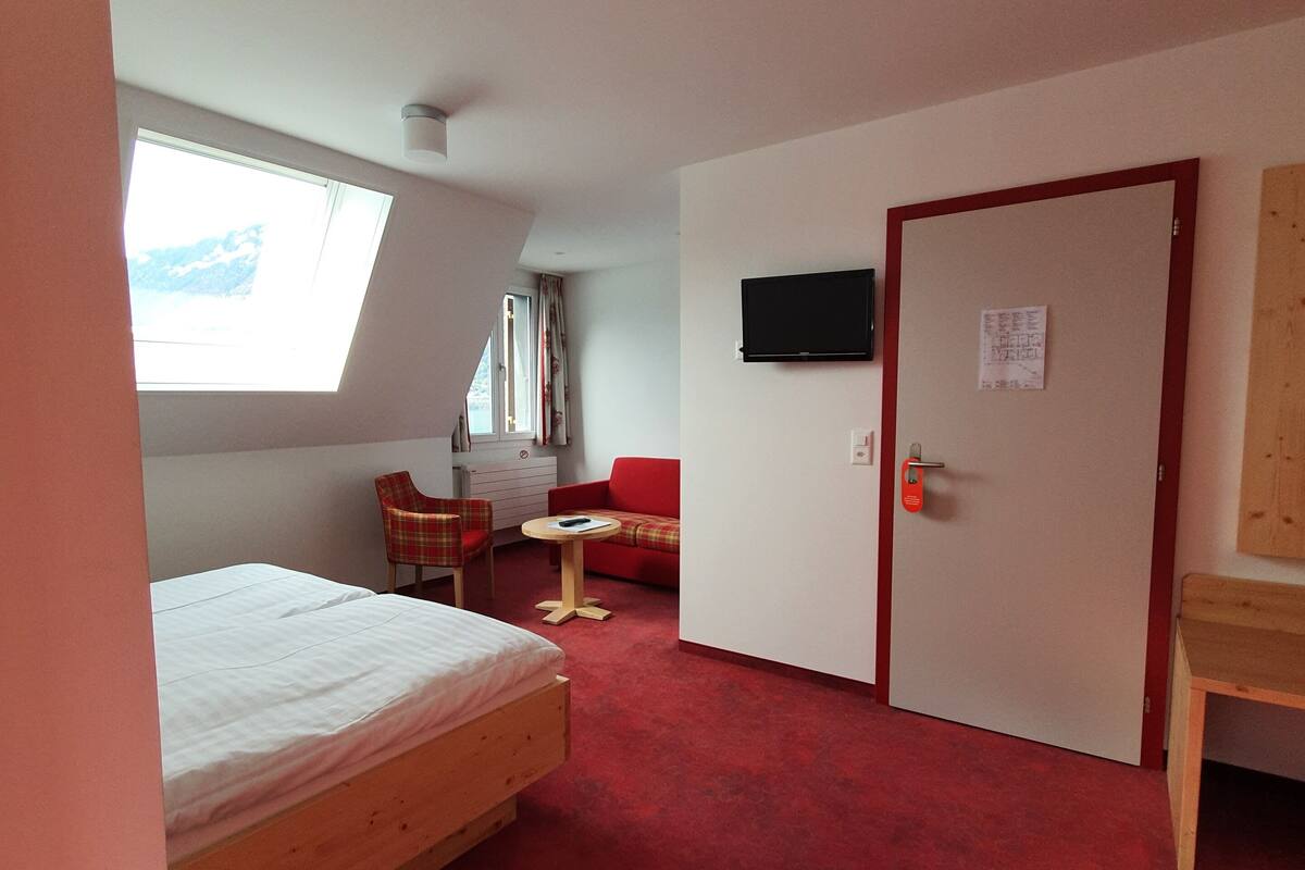 A room with a bed, a red sofa, a small table, a TV mounted on the wall, and a door with a sign.