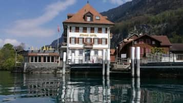 A hotel with a traditional European architectural style, featuring a prominent central building with a red roof and multiple windows. It is situated on stilts over a body of water, reflecting the building and the surrounding landscape.