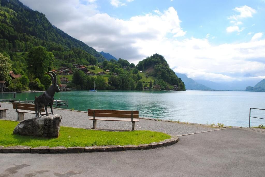 A park with a bench and a sculpture of a goat by a lake, surrounded by mountains and houses.
