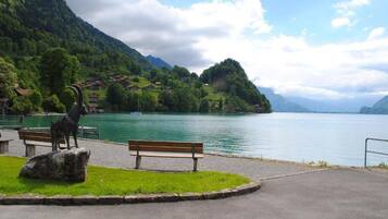 A park with a bench and a sculpture of a goat by a lake, surrounded by mountains and houses.