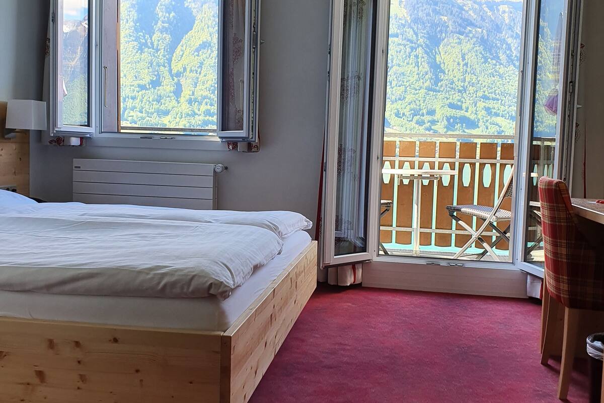 A bedroom with a wooden bed, a red carpet, and a balcony with a view of mountains.